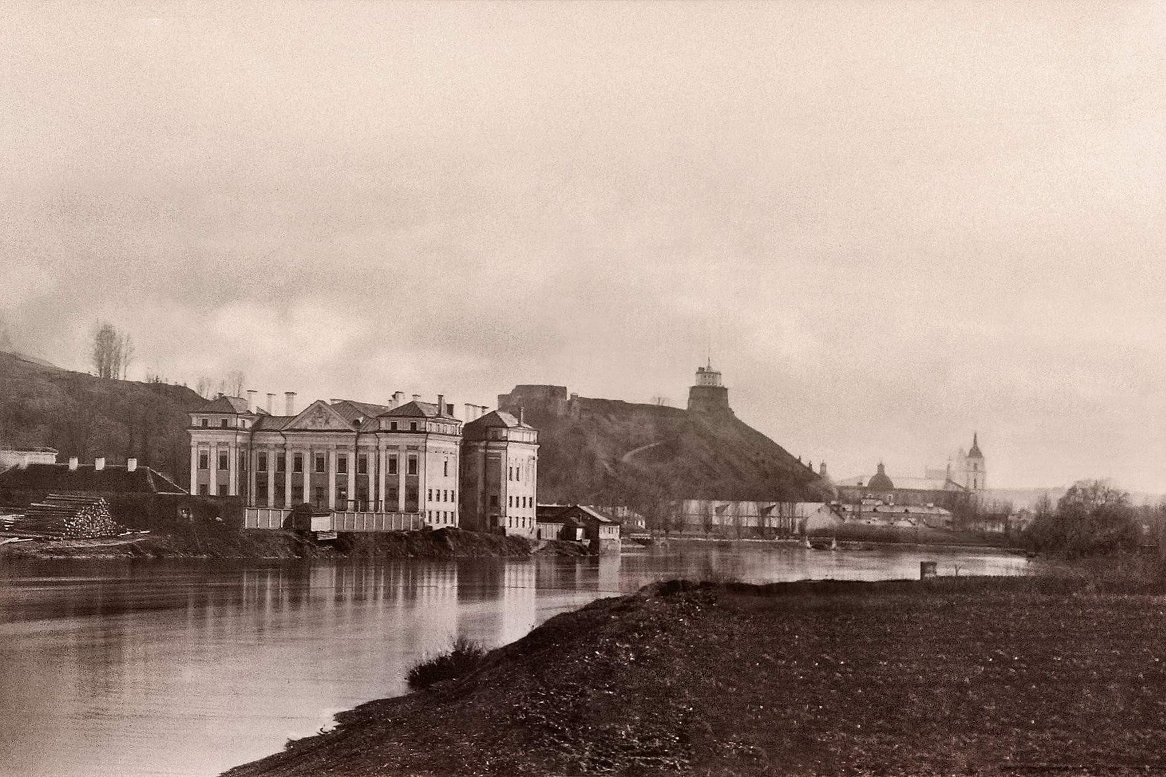 Istorinė Sluškų rūmų Vilniuje nuotrauka. Rūmai stovi Neries pakrantėje, fone matyti Pilies kalnas su Gedimino bokštu ir miesto panorama.
