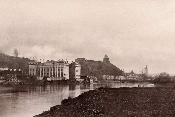 Istorinė Sluškų rūmų Vilniuje nuotrauka. Rūmai stovi Neries pakrantėje, fone matyti Pilies kalnas su Gedimino bokštu ir miesto panorama.