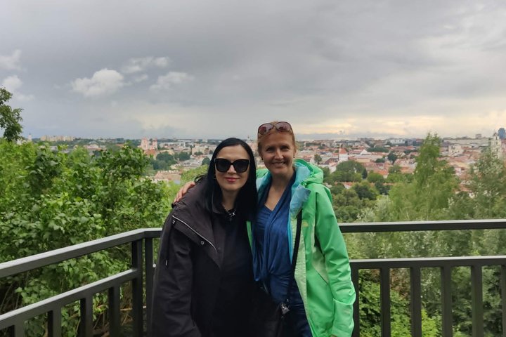 Dvi moterys stovi Vilniaus pilių rezervato apžvalgos bokštelyje, už jų atsiveria miesto senamiesčio panorama, žaliuojantys medžiai ir debesuotas dangus.