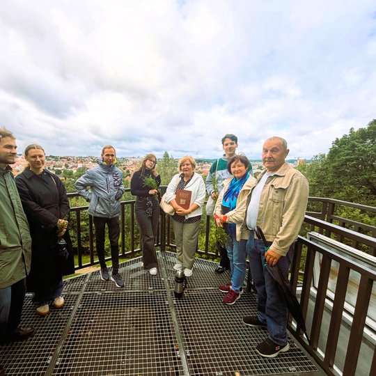 Ekskursijos dalyviai Altanos kalno apžvalgos aikštelėje Vilniuje. Grupė pozuoja nuotraukai, o fone atsiveria miesto panorama ir debesuotas dangus.