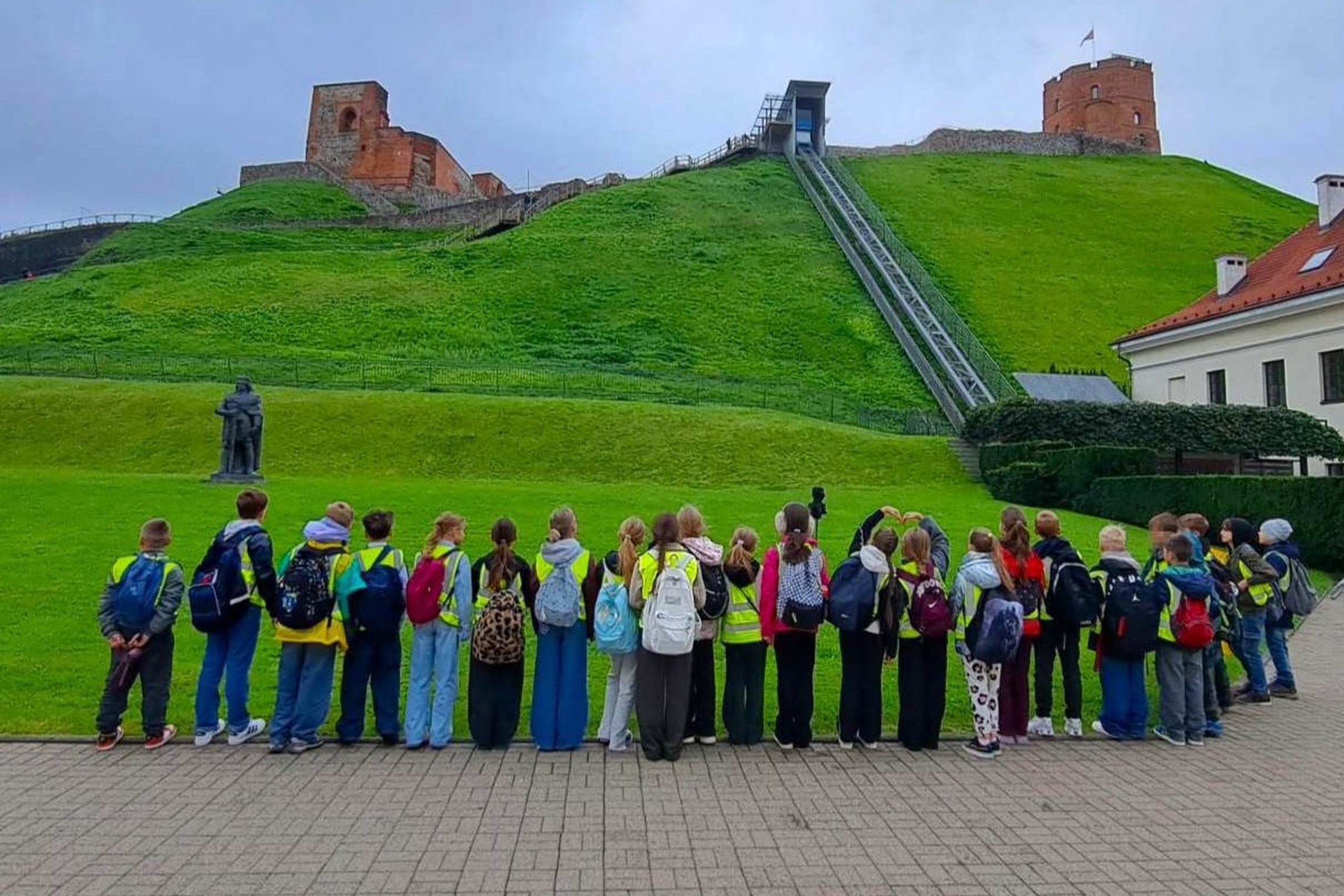 Moksleivių grupė Vilniaus pilių teritorijoje dalyvauja edukacijoje „Gediminas ir Vilniaus trys pilys“. Fone matyti Gedimino kalnas, Aukštutinės pilies liekanos ir Gedimino pilies bokštas.