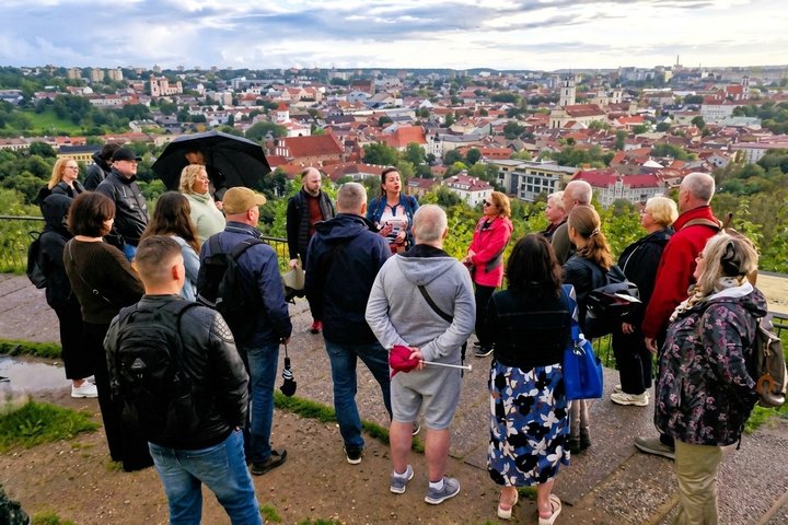 Ekskursijos dalyvių grupė klausosi gidės ant Vilniaus pilių kalvų, fone atsiveria plati Senamiesčio panorama su raudonais stogais ir bažnyčių bokštais.