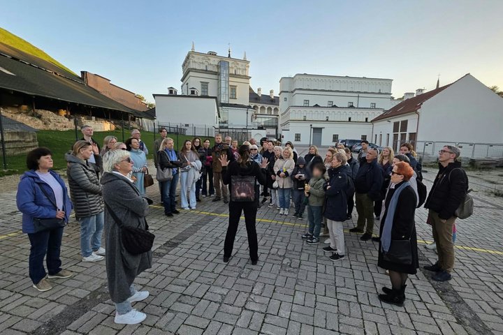 Grupė ekskursijos dalyvių stovi Vilniaus pilių teritorijoje prie Valdovų rūmų ir klausosi gidės pasakojimo.