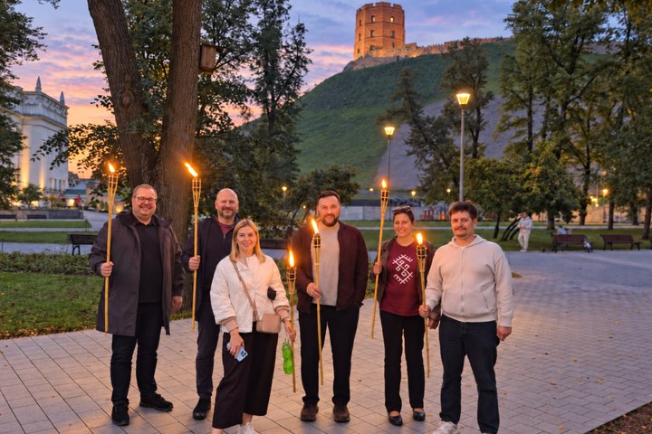 Šeši žmonės su deglais pozuoja parke Vilniuje, Gedimino pilies bokšto papėdėje, rausvo vakaro dangaus fone.