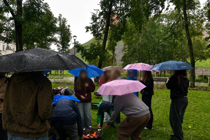 Mokiniai po skėčiais lietingą dieną prie laužo su skėčiais kepa zefyrus Vilniaus pilių rezervate.