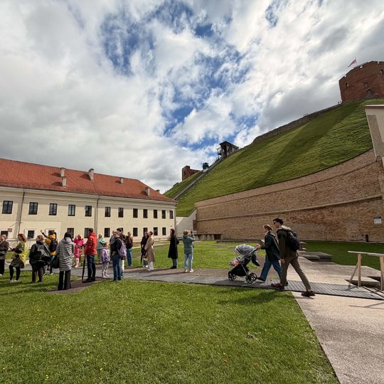 Ekskursijos dalyviai Vilniaus pilių teritorijoje prie Gedimino kalno. Fone matyti žaliuojantis kalno šlaitas, Gedimino pilies bokštas ir debesuotas, protarpiais pragiedrėjantis dangus.
