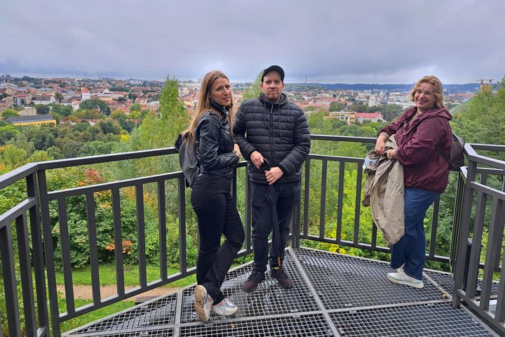Trys ekskursijos dalyviai stovi apžvalgos aikštelėje, nuo kurios atsiveria plati Vilniaus senamiesčio panorama. Fone matyti miesto pastatai, bažnyčių bokštai ir žaliuojanti Vilniaus pilių teritorija.