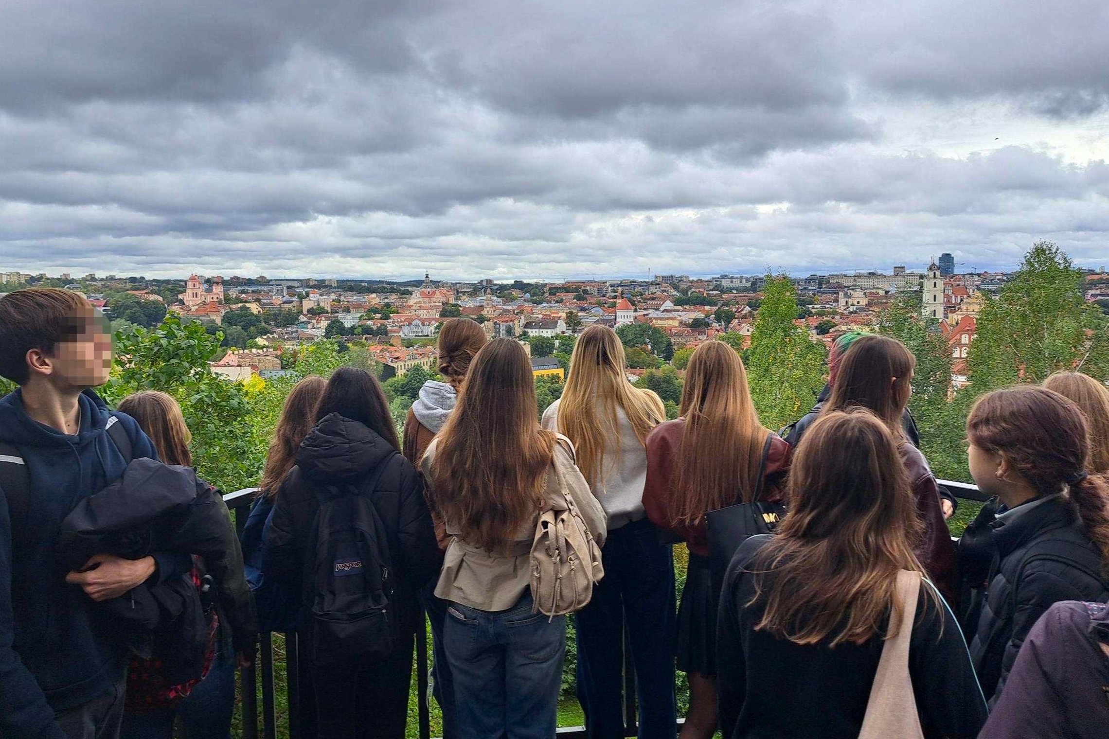 Nuotraukoje grupė Vilniaus Užupio gimnazijos moksleivių stovi apžvalgos aikštelėje ir žvelgia į Vilniaus senamiesčio panoramą. Fone matyti miesto stogai, bažnyčių bokštai ir debesuotas dangus.