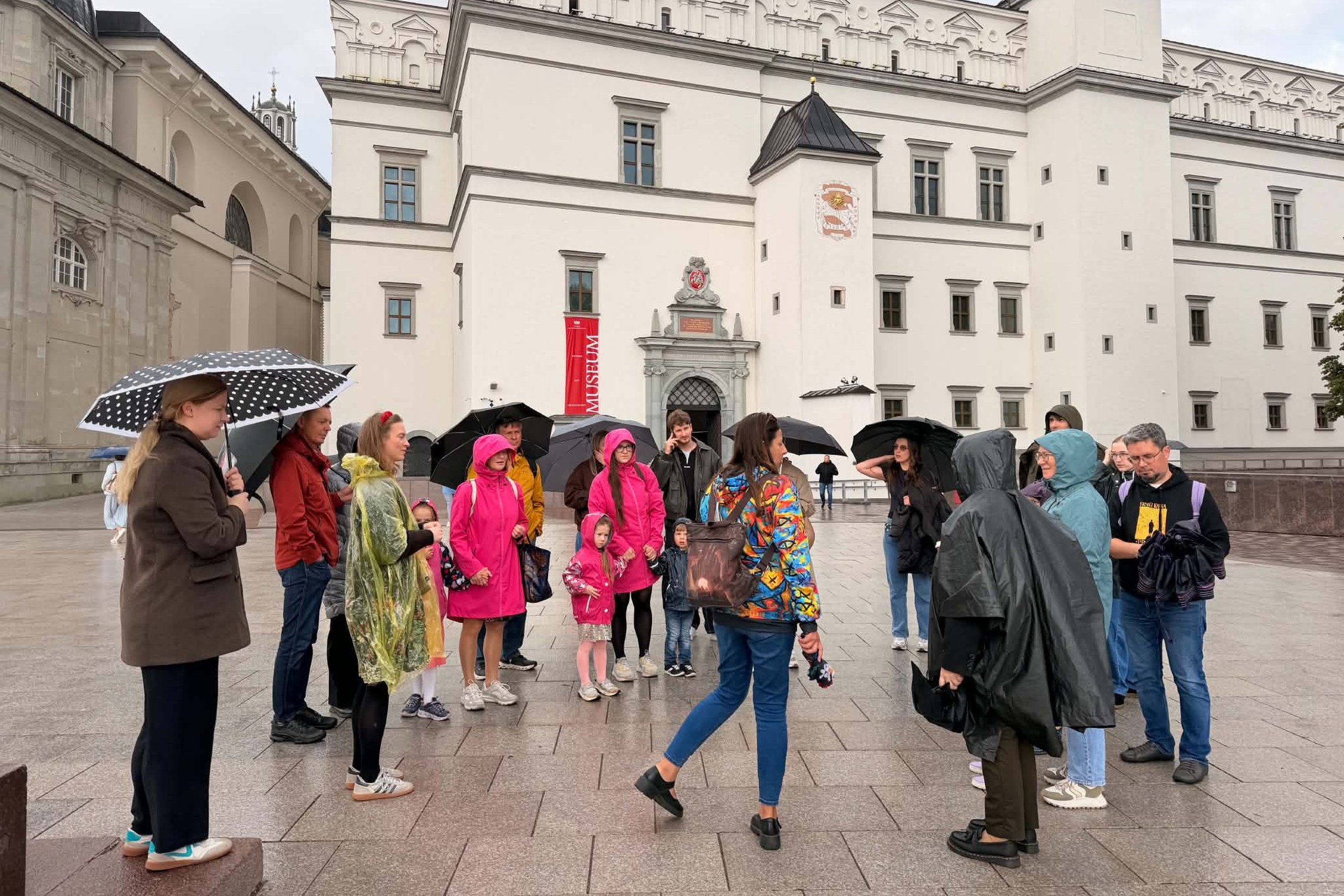 Ekskursijos dalyviai lietingą ir vėjuotą dieną stovi Žemutinės pilies teritorijoje Vilniuje, prie Valdovų rūmų.
