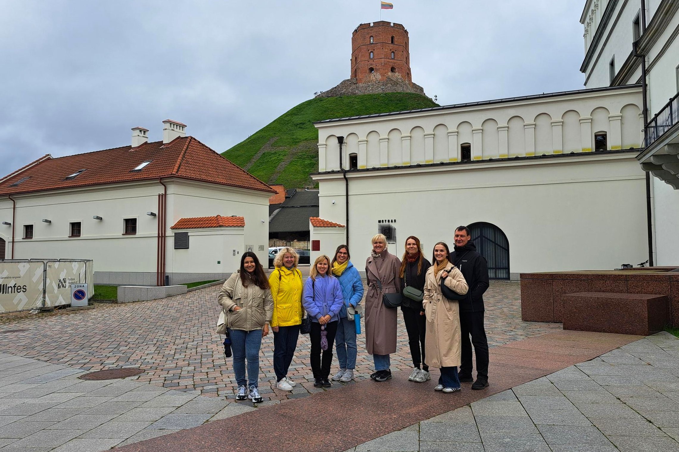Grupė ekskursijos dalyvių pozuoja Vilniaus Žemutinės pilies teritorijoje. Fone matyti Gedimino kalnas su Aukštutinės pilies bokštu ir Lietuvos vėliava.